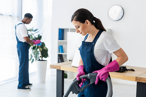 Selective focus of attractive worker of cleaning service using vacuum cleaner near african american colleague in office