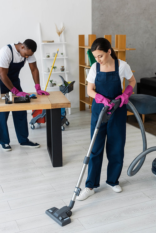 Selective focus of attractive cleaner vacuuming floor near african american colleague with rag in office