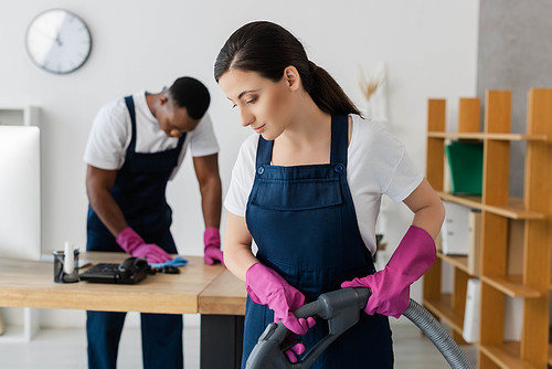 Selective focus of cleaner in overalls and rubber gloves using vacuum cleaner near african american colleague cleaning table in office