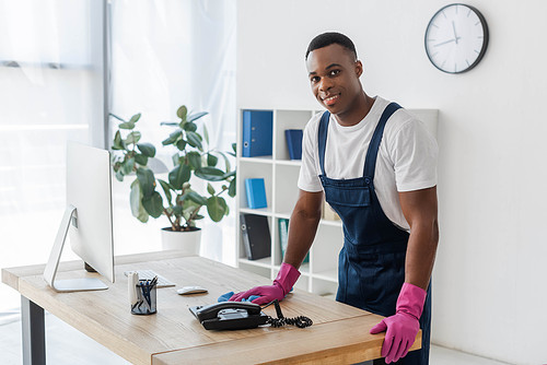 Smiling african american cleaner with rag standing near office table