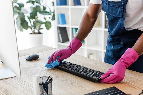 Cropped view of african american cleaner in rubber gloves cleaning computer keyboard on office table