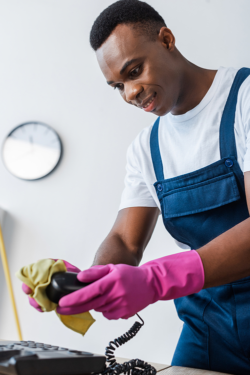 Selective focus of smiling african american cleaner rubbing telephone on office table