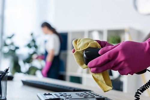 Selective focus of cleaner in rubber gloves cleaning telephone near colleague in office