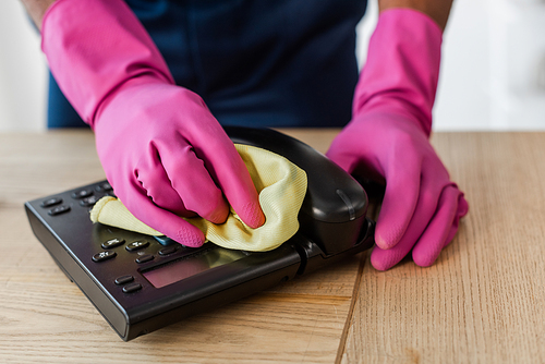 Cropped view of african american cleaner in rubber gloves cleaning telephone with rag on table in office