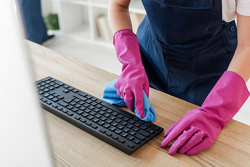 Cropped view of cleaner in rubber gloves holding rag near computer keyboard in office