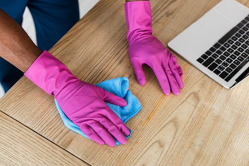 Cropped view of african american cleaner in rubber gloves cleaning table near laptop in office