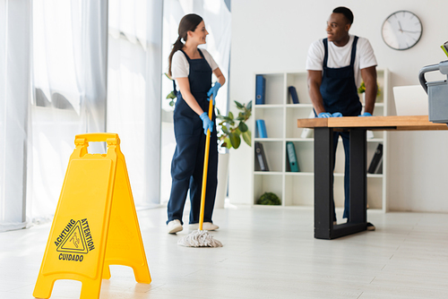 Selective focus of wet floor sign and smiling multiethnic cleaners working in office