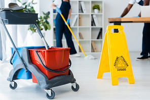 Selective focus of cleaning trolley near wet floor sign and multiethnic cleaners working in office