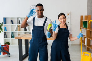 Multicultural cleaners with cleaning supplies smiling at camera and showing okay gesture in office