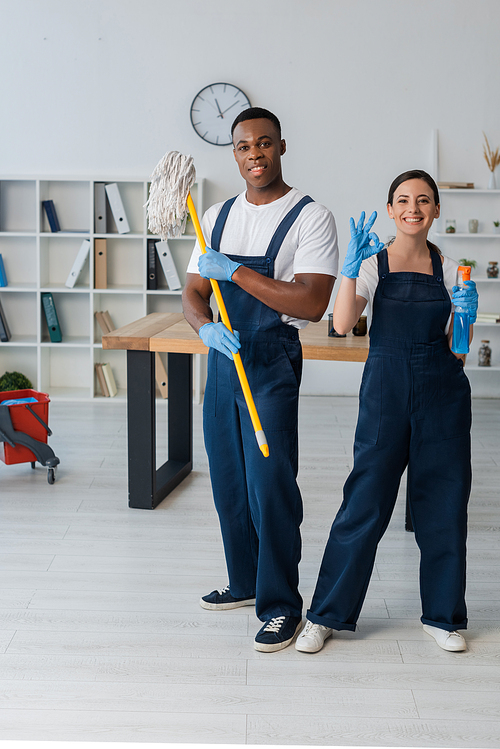 Smiling multiethnic cleaners holding mop and detergent and showing ok gesture in office