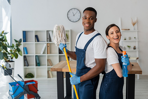 Positive multiethnic cleaners holding mop and detergent while smiling at camera in office