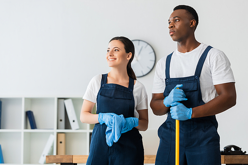 Smiling cleaner holding rag near african american colleague with mop in office