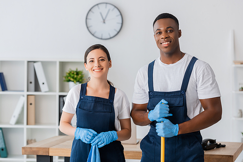 Smiling multiethnic workers of cleaning service holding rag and mop while working in office