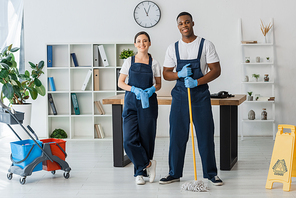 Multicultural cleaners with rag and mop smiling at camera near wet floor sign and cleaning trolley in office