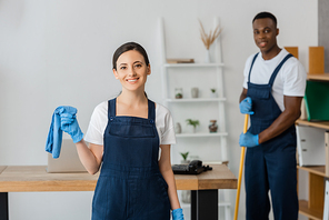 Selective focus of smiling cleaner in uniform holding rag near african american colleague washing floor in office