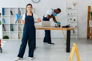 Selective focus of smiling cleaner holding rag near african american colleague washing floor in office