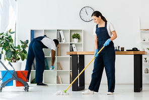 Selective focus of smiling cleaner washing floor with mop near african american colleague in office