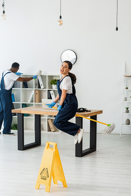 Selective focus of positive cleaner jumping with mop near african american colleague working in office