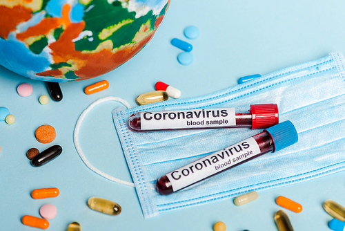 test tubes with blood samples and coronavirus lettering near medical mask, pills and globe on blue background