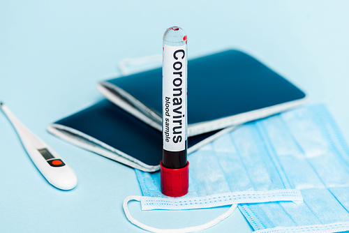 selective focus of test tube with blood sample and coronavirus lettering near medical masks, thermometer and passports on blue background