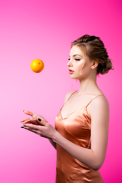 side view of elegant beautiful blonde woman throwing ripe orange isolated on pink