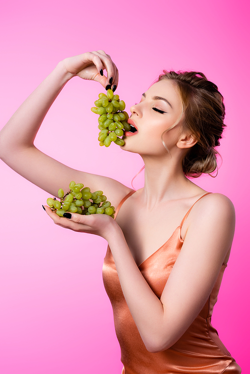 elegant beautiful blonde woman eating green grapes isolated on pink