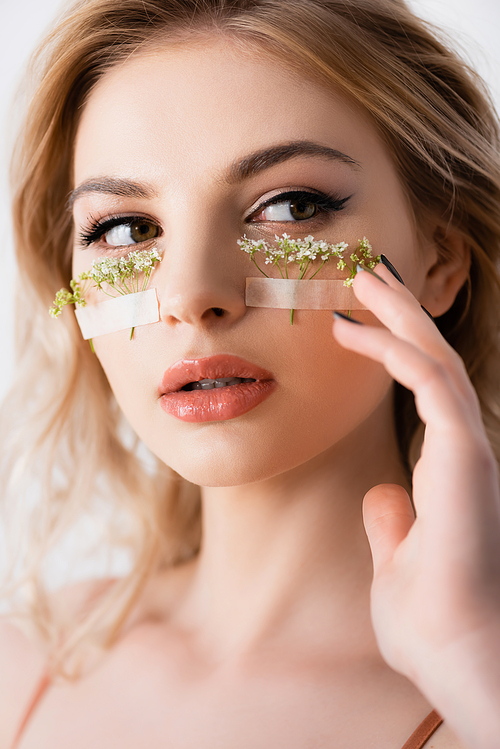 beautiful blonde woman touching face with wildflowers under eyes isolated on white