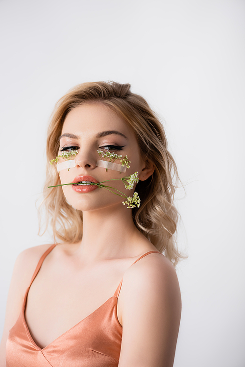 beautiful blonde woman in silk dress with wildflowers under eyes and in mouth isolated on white