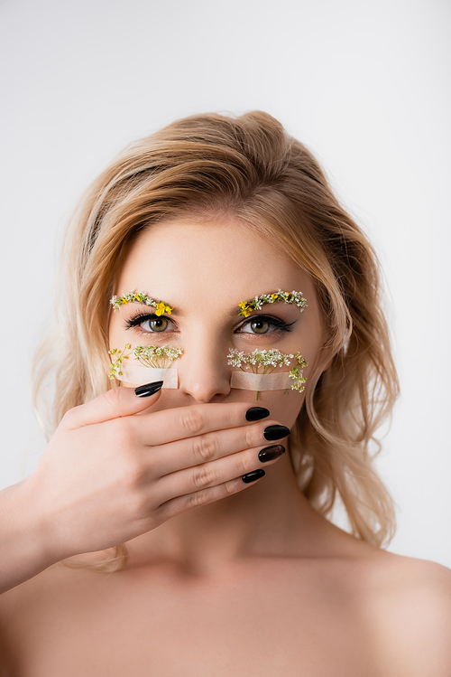 naked beautiful blonde woman with wildflowers under eyes covering mouth isolated on white