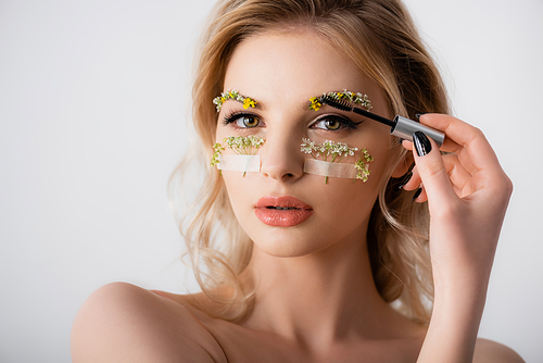 naked beautiful blonde woman styling wildflowers on eyebrows with brow gel isolated on white