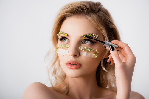 naked beautiful blonde woman styling wildflowers on eyebrows with brow gel isolated on white