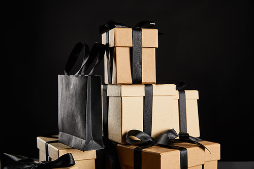 stack of cardboard gift boxes with black ribbons and shopping bag isolated on black, black Friday concept