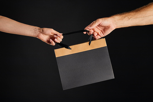 cropped view of woman and man holding paper shopping bag isolated on black, black Friday concept