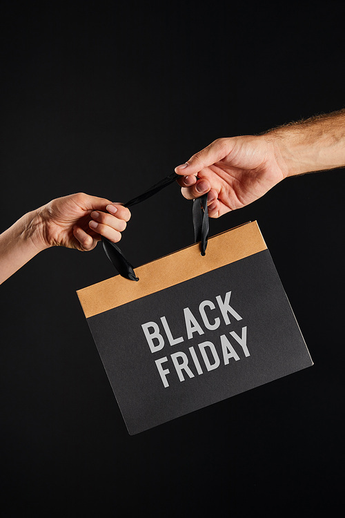 cropped view of woman and man holding paper shopping bag with black Friday lettering isolated on black