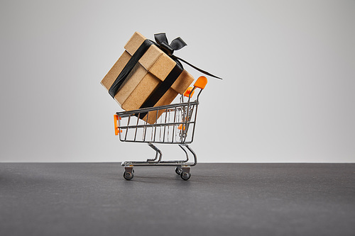 cardboard gift box with black ribbon in small shopping cart isolated on grey, black Friday concept