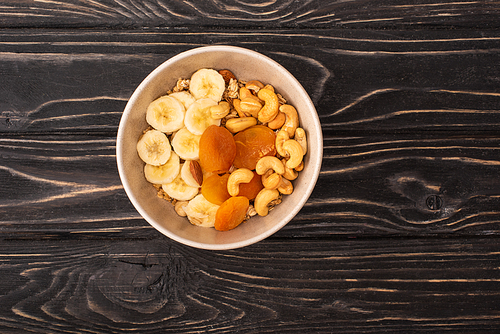 top view of delicious granola with nuts, banana and dried apricots on wooden black surface