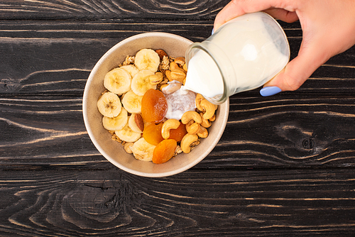 cropped view of woman adding yogurt to delicious granola with nuts, banana and dried apricots on wooden black surface