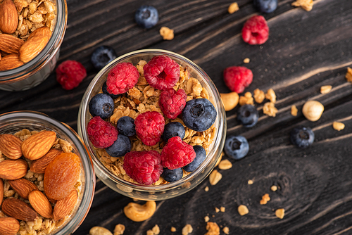 top view of delicious granola with berries, dried apricots, nuts and yogurt in glass cups on wooden surface