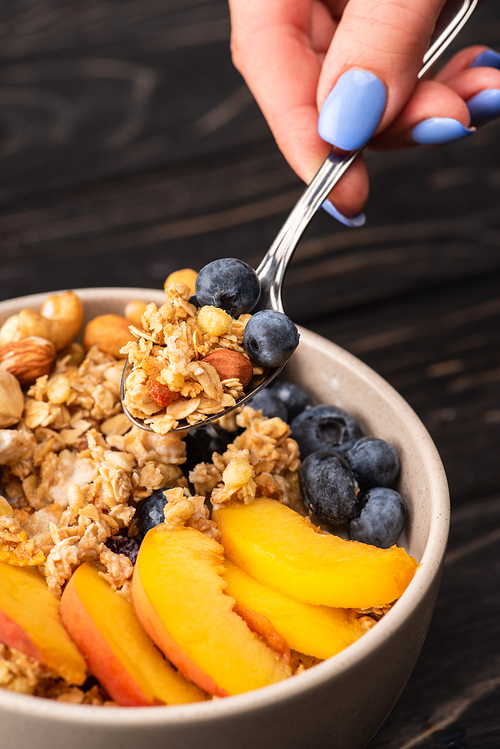 cropped view of woman eating delicious granola with nuts, peach, blueberry