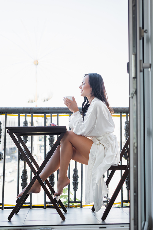 smiling brunette woman in white robe drinking tea and sitting on balcony