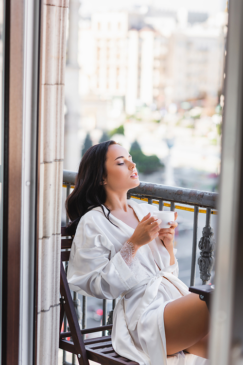 beautiful brunette woman in white robe drinking coffee on balcony