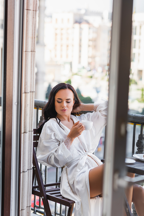 beautiful brunette woman in white robe drinking coffee on balcony