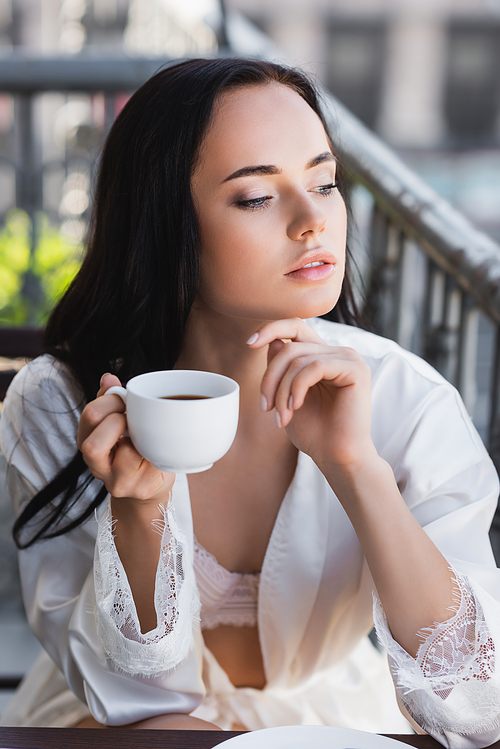 beautiful brunette woman in white robe drinking coffee on balcony