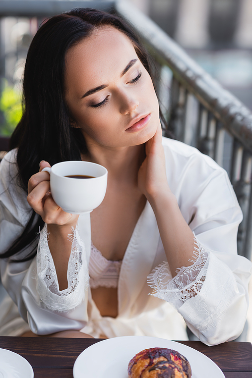 brunette woman in white robe drinking coffee and touching neck on balcony