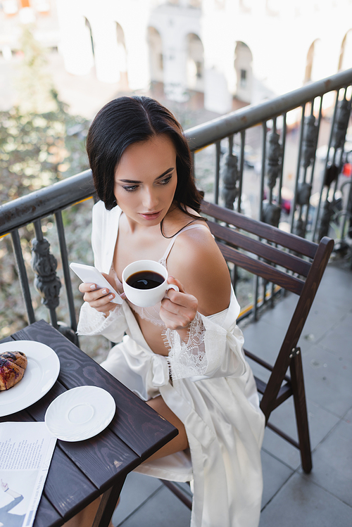 beautiful brunette woman in white robe holding smartphone and drinking coffee on balcony