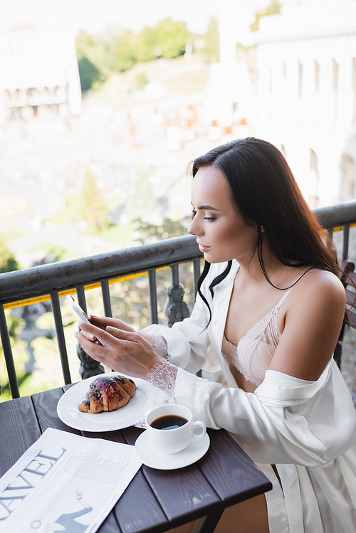 brunette woman having breakfast on balcony and texting on smartphone
