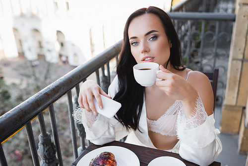 beautiful brunette woman in white robe drinking coffee on balcony and holding smartphone