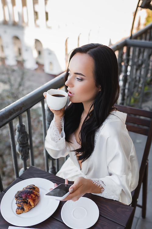 beautiful brunette woman in white robe drinking coffee and looking away