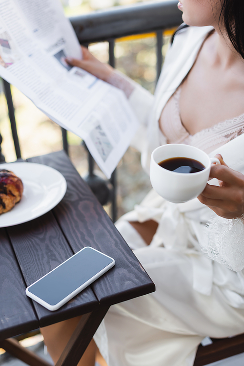partial view of brunette woman in white robe drinking coffee and reading newspaper