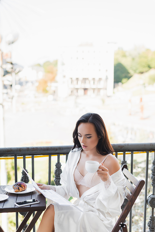 brunette woman in white robe sitting on balcony and reading newspaper